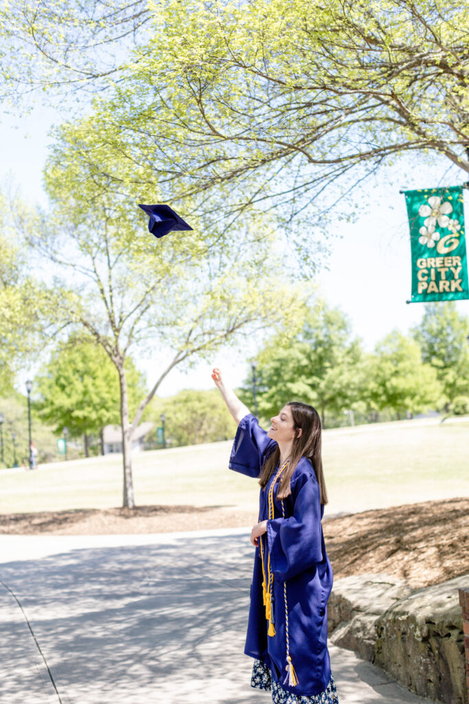 A senior girl in her cap and gown tossing her cap in the air during a Greenville SC senior session with Jenny Macy Photography at Greer City Park.