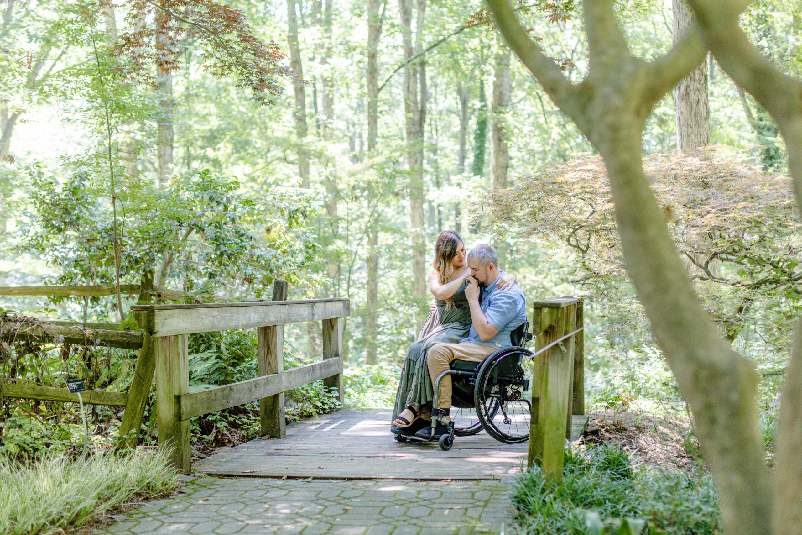 A woman sitting on her fiance's lap in a wheelchair while he kisses her hand at Clemson Botanical Gardens in Greenville SC