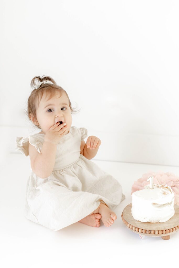A baby in a neutral dress in a natural light white wall studio with a smash cake.