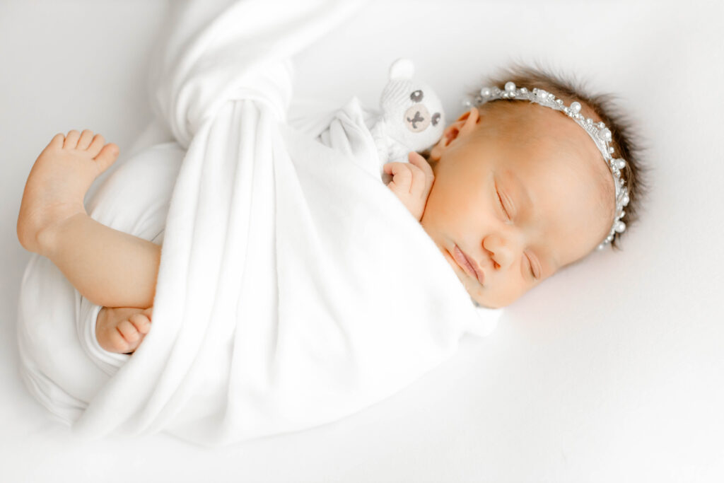 A sleeping baby swaddled in a white blanket holding a small teddy bear during a newborn photo session in greenville, SC